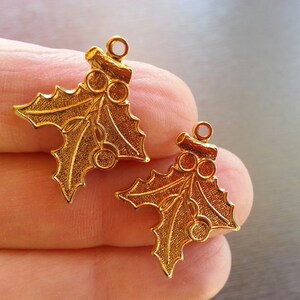 May include: Two gold-tone metal holly leaf charms with a loop at the top for hanging. Each charm features a detailed holly leaf design with three red berries.