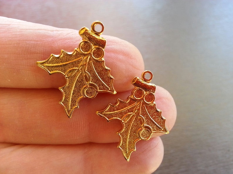 May include: Two gold-tone metal holly leaf charms with a loop at the top for hanging. Each charm features a detailed holly leaf design with three red berries.