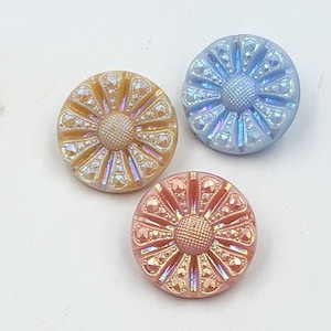May include: Three iridescent buttons with a flower design. The buttons are in shades of blue, pink, and gold.