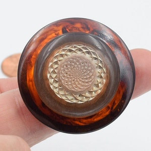 May include: A large, round button with a tortoiseshell-patterned rim and a clear, textured center. The center features a detailed, spiral-like design. The button is a mix of brown, amber, and clear colours.