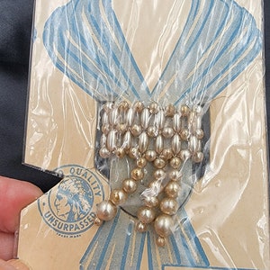 May include: A vintage package for a decorative scarf slide. The package features a drawing of a woman wearing a scarf and the text "Scarf Slide". The slide is made of gold and white beads and is packaged in a clear plastic bag. The package also includes the text "Quality Unsurpassed" and "Made in Japan".