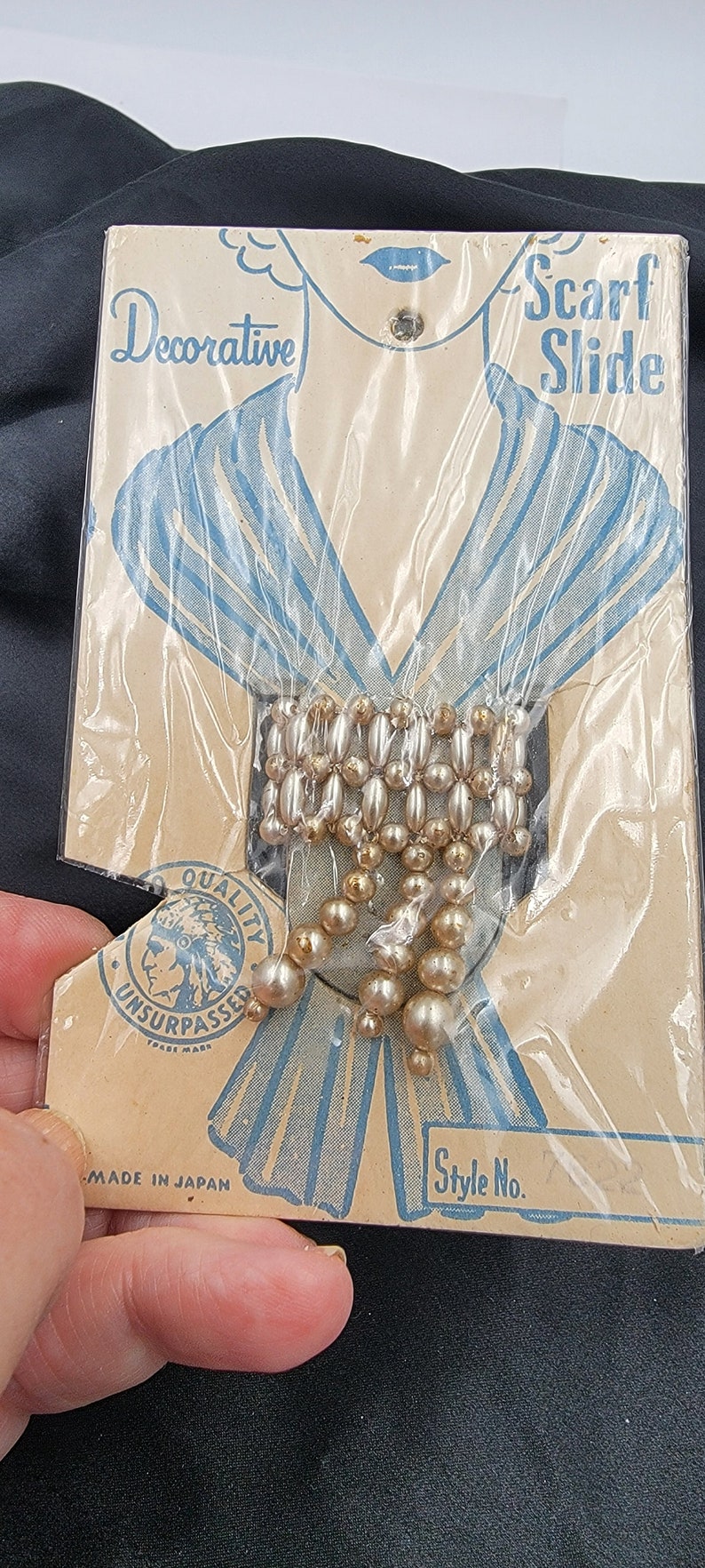 May include: A vintage package for a decorative scarf slide. The package features a drawing of a woman wearing a scarf and the text "Scarf Slide". The slide is made of gold and white beads and is packaged in a clear plastic bag. The package also includes the text "Quality Unsurpassed" and "Made in Japan".