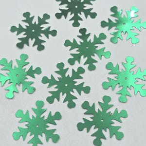 May include: Nine green metallic snowflake sequins. The sequins are about 1 inch in diameter and have a hole in the center.