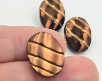 May include: Three oval-shaped buttons with a copper and gold striped design. The buttons have a glossy finish and are set against a white background. The buttons are likely for sewing or crafts.