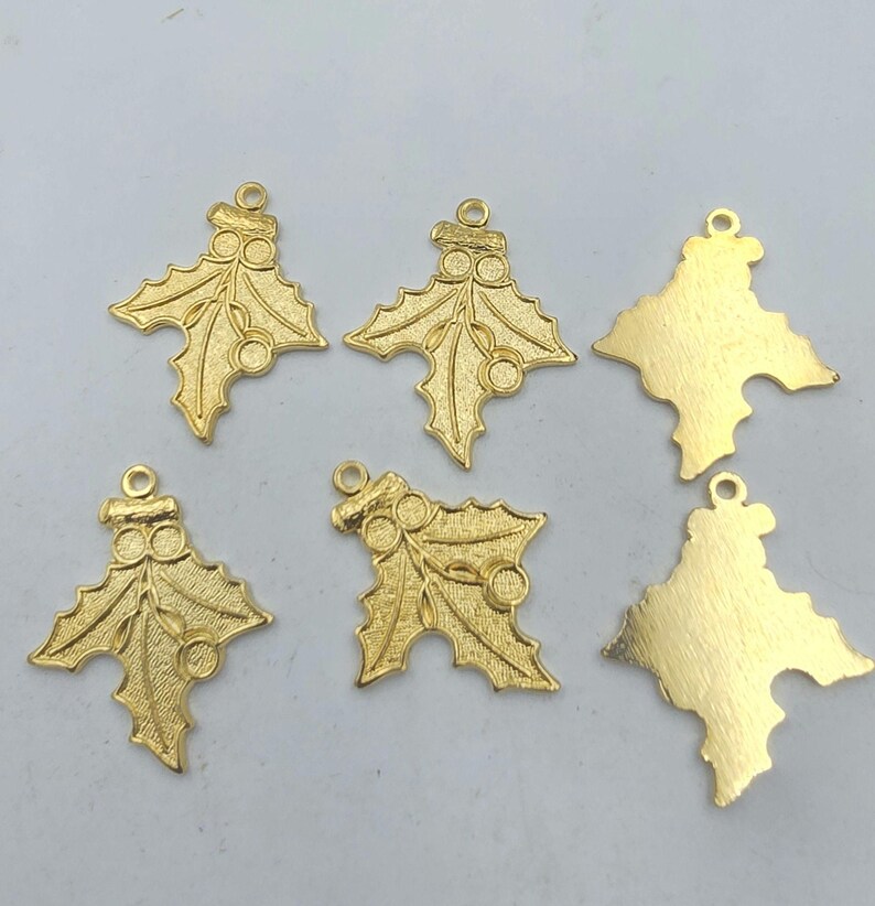 May include: Six gold-colored metal charms in the shape of holly leaves with red berries. The charms have a loop at the top for attaching to a necklace or bracelet.