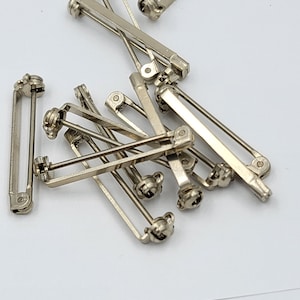 May include: A collection of silver-colored brooch backs, scattered on a white surface. The brooch backs have a rectangular shape with a pin and a clasp mechanism. They are likely used for attaching pins or badges to clothing or fabric.