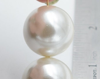 Puede incluir: Dos grandes cuentas redondas de perlas blancas están suspendidas de un alambre dorado. Las cuentas tienen aproximadamente 2,5 cm de diámetro. Una regla en el fondo muestra la medida en pulgadas. Las cuentas son lisas y brillantes.