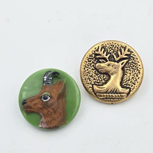 Könnte beinhalten: Zwei runde Knöpfe mit einem Hirsch-Motiv. Ein Knopf ist grün mit einem braunen Hirschkopf, der andere ist golden mit einem Hirschkopf und Geweih.