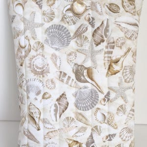 May include: A rectangular, quilted pillow cover with a repeating pattern of seashells and starfish in beige, brown, and grey on a white background. A gold-coloured seashell charm is attached to the top.