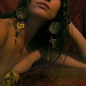 May include: A woman wearing long, dark hair with two large, ornate earrings. The earrings are made of gold metal and have a variety of charms, including a bull's head, a small green stone, and a round disc with a gold center.