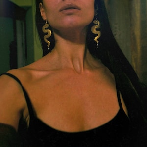 May include: A woman wearing a black tank top with gold snake earrings. The earrings are long and dangle from her ears.
