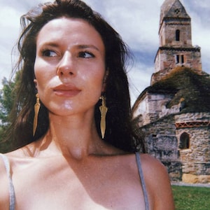 May include: A woman with long dark hair wearing a grey tank top and gold dagger earrings stands in front of a stone building with a tower.