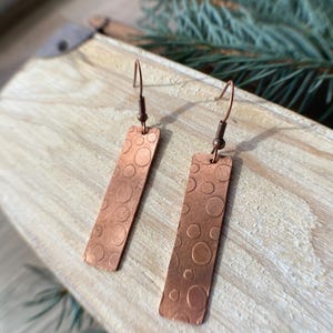 May include: A pair of copper earrings with a hammered texture and a circle pattern. The earrings are hanging from a wooden surface.