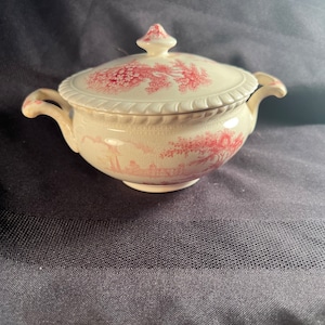 Puede incluir: Un azucarero de cerámica blanca con tapa, con un diseño floral y de paisaje rosa. El tazón tiene dos asas y una perilla en la tapa.