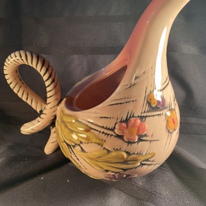 May include: A pink ceramic jug with a floral design. The jug has a twisted handle and a spout that curves upwards. The flowers are in shades of pink, yellow, and purple.