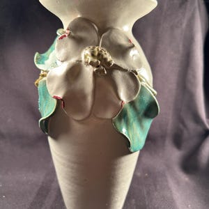 May include: A handcrafted ceramic vase with a cream-colored base and a flared top. The vase is adorned with a large, three-dimensional flower with white petals, green leaves, and red accents. The vase is approximately 10 inches tall.