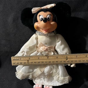 Pu&ograve; includere: Un abito bianco di raso con bordino di pizzo e un fiocco rosa. L'abito &egrave; indossato da una bambola di peluche Minnie Mouse con orecchie nere e un fiocco rosa. La bambola indossa scarpe rosa ed &egrave; alta circa 25 cm.
