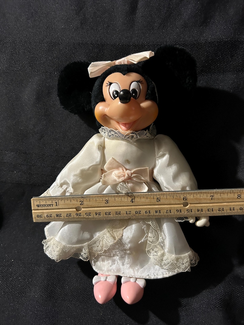 Pu&ograve; includere: Un abito bianco di raso con bordino di pizzo e un fiocco rosa. L'abito &egrave; indossato da una bambola di peluche Minnie Mouse con orecchie nere e un fiocco rosa. La bambola indossa scarpe rosa ed &egrave; alta circa 25 cm.