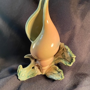 May include: A ceramic seashell-shaped vase in shades of tan, pink, and teal. The vase has a smooth, glossy finish and is designed with a flared opening. The base features a detailed, textured design, adding to its unique aesthetic.