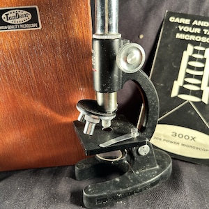 May include: A black Tasco Deluxe microscope with a silver eyepiece and objective lenses, featuring a stage with metal clips. It is accompanied by a wooden storage case with a metal label and a manual titled Care and Your Tasco Microscope 300X.
