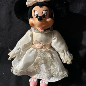 Pu&ograve; includere: Una bambola di peluche vintage di Minnie Mouse che indossa un abito di raso bianco con bordino in pizzo e un fiocco di raso rosa. Ha le orecchie nere e un fiocco rosa sulla testa.