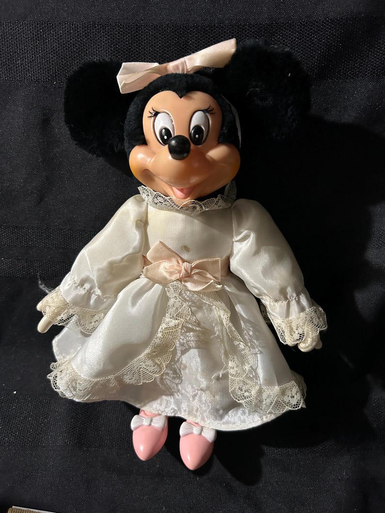 Pu&ograve; includere: Una bambola di peluche vintage di Minnie Mouse che indossa un abito di raso bianco con bordino in pizzo e un fiocco di raso rosa. Ha le orecchie nere e un fiocco rosa sulla testa.