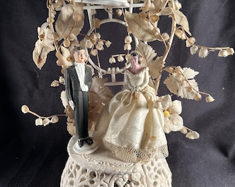 Op de afbeelding: Een bruidstaarttopper met een bruid in een crèmekleurige jurk en een bruidegom in een zwart smoking op een witte, sierlijke, ronde voet. Daarachter staat een wit draadframe versierd met gedroogde witte bloemen en een witte bel met een lint.
