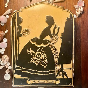 Könnte beinhalten: Ein Vintage-Metallschild mit einer schwarzen Silhouette einer Frau in einem Kleid und einem Mann, der hinter ihr steht. Das Schild ist cremefarben mit goldenen Akzenten und dem Text "The Final Touch" in einem Banner unten.