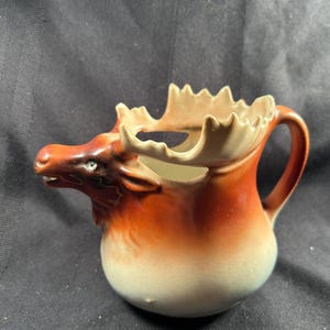 Könnte beinhalten: Eine Keramik-Sahnekännchen in Form eines Elchs. Das Kännchen ist überwiegend braun mit einem weißen Sockel und hellbraunen Geweih. Es hat einen Griff und einen Ausguss in Form der Schnauze des Elchs.