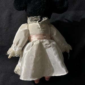 Pu&ograve; includere: Una bambola di porcellana bianca e nera che indossa un vestito bianco con bordi in pizzo e una cintura rosa. La bambola ha una testa di peluche nero con un fiocco.