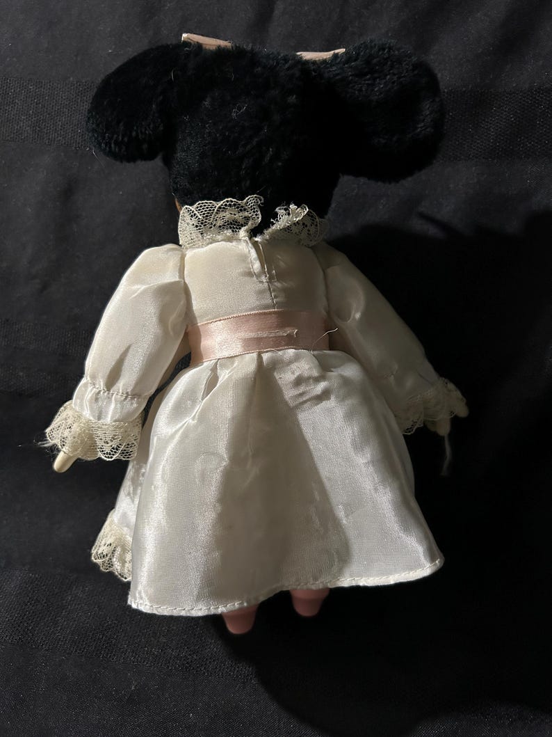 Pu&ograve; includere: Una bambola di porcellana bianca e nera che indossa un vestito bianco con bordi in pizzo e una cintura rosa. La bambola ha una testa di peluche nero con un fiocco.