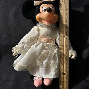 Pu&ograve; includere: Una bambola di peluche di Minnie Mouse che indossa un vestito bianco con pizzo e un fiocco rosa. Ha le orecchie nere e un fiocco rosa nei capelli. Indossa scarpe rosa.