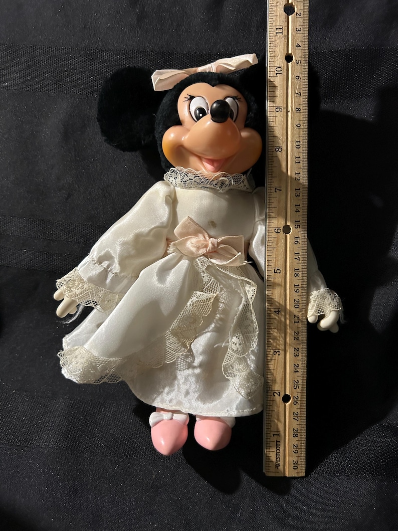 Pu&ograve; includere: Una bambola di peluche di Minnie Mouse che indossa un vestito bianco con pizzo e un fiocco rosa. Ha le orecchie nere e un fiocco rosa nei capelli. Indossa scarpe rosa.