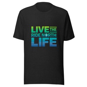 以下が含まれることがあります： 「LIVE THE RIDE NORTH LIFE」の文字が緑と青のグラデーションでプリントされたダークグレーのTシャツ。デザインにはコンパスのシンボルが含まれています。クルーネックのTシャツです。