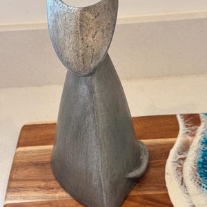 May include: A silver-toned, stylised cat figurine with a smooth, tapered body and pointed ears. The sculpture features a textured, metallic finish and is positioned on a wooden board with a blue and white resin wave design along the edge.