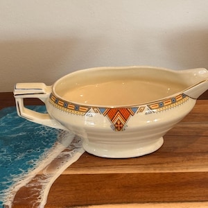May include: A cream-coloured ceramic gravy boat featuring a geometric Art Deco pattern in orange, blue, and gold along the rim. The vessel has a wide pouring spout, a sturdy angular handle, and a pedestal base, showcasing a smooth, glossy finish.