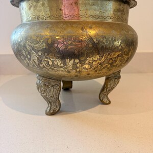 May include: A round brass bowl with a wide rim, featuring intricate etched floral patterns across the body and a smooth upper band. The vessel is supported by three ornate, curved legs that display detailed relief carvings.