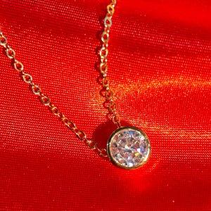 Puede incluir: Un collar de cadena dorada con un colgante redondo que presenta una piedra clara y brillante.