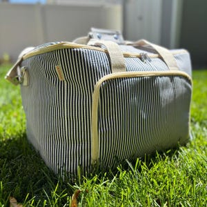 May include: A large, rectangular tote bag with a black and white striped pattern. The bag has beige trim, handles, and a zipper closure. The bag is sitting on green grass.