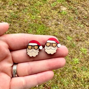 May include: Two wooden Santa Claus earrings. Each earring features a red hat, round glasses, and a white beard. The earrings are held in a hand against a background of green grass.