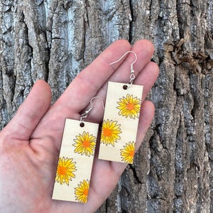 May include: A pair of wooden earrings with a yellow and orange floral pattern. The earrings are rectangular in shape and have silver hooks.