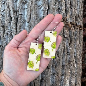 May include: A pair of wooden earrings with a white background and a green tropical leaf pattern. The earrings are rectangular in shape and have silver hooks.