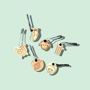 May include: Six wooden stitch markers with different designs, including a sock, a sheep, a ball of yarn, and a knitted fabric. Each marker has a metal loop for attaching to yarn.