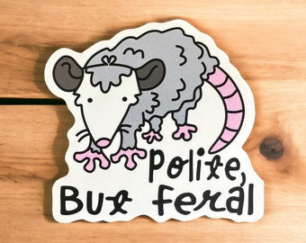 May include: A white sticker with a cartoon illustration of a gray opossum with pink ears, nose, and tail. The opossum is standing on its hind legs with its front paws outstretched. The text "polite, but feral" is written below the opossum.