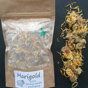 Puede incluir: Una bolsa de papel marr&oacute;n con una ventana de pl&aacute;stico transparente que contiene p&eacute;talos de cal&eacute;ndula secos amarillos y naranjas. Una etiqueta blanca dice Marigold 100% biodegradable packaging con un dibujo de una oveja. Flores secas sueltas al lado.