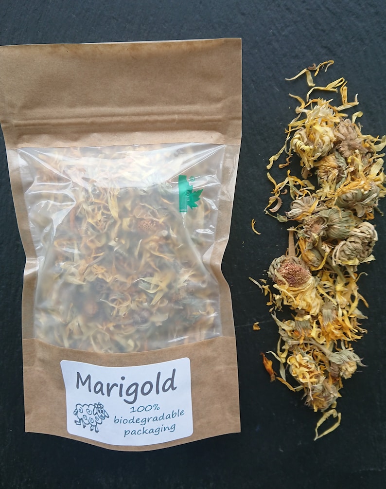 Puede incluir: Una bolsa de papel marr&oacute;n con una ventana de pl&aacute;stico transparente que contiene p&eacute;talos de cal&eacute;ndula secos amarillos y naranjas. Una etiqueta blanca dice Marigold 100% biodegradable packaging con un dibujo de una oveja. Flores secas sueltas al lado.