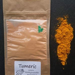 Puede incluir: Una bolsa de papel marr&oacute;n con una ventana transparente que contiene c&uacute;rcuma en polvo, con una etiqueta blanca con el texto Tumeric, un dibujo de una oveja y las palabras 100% biodegradable packaging. Un mont&oacute;n de polvo amarillo brillante al lado.
