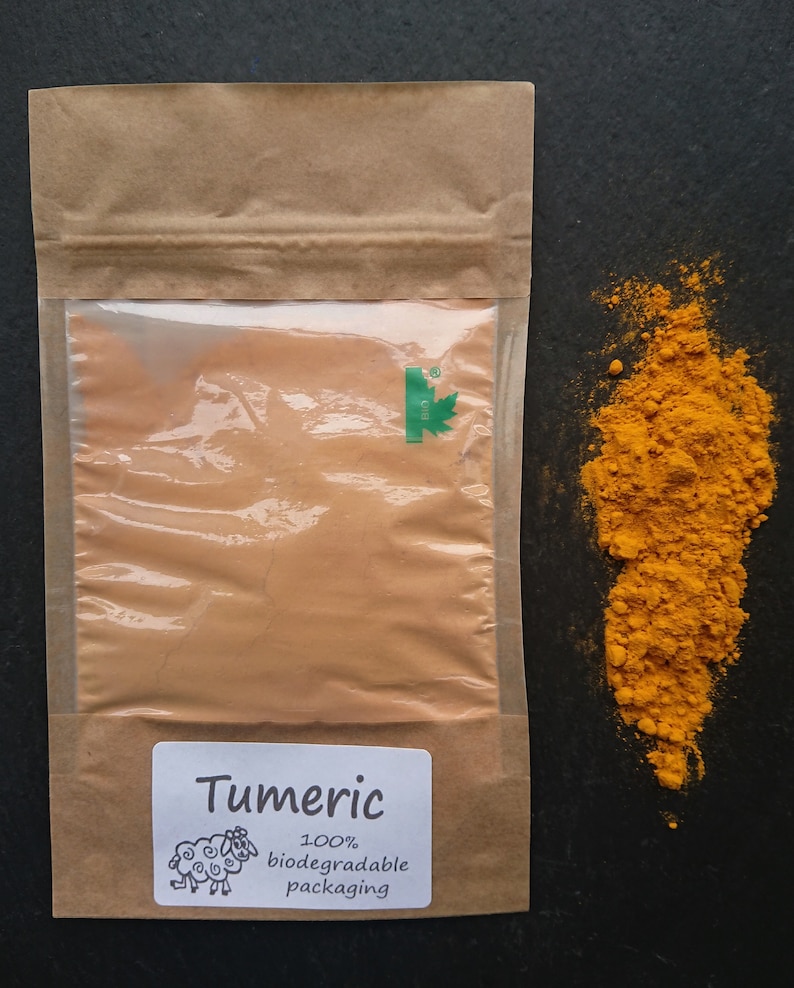 Puede incluir: Una bolsa de papel marr&oacute;n con una ventana transparente que contiene c&uacute;rcuma en polvo, con una etiqueta blanca con el texto Tumeric, un dibujo de una oveja y las palabras 100% biodegradable packaging. Un mont&oacute;n de polvo amarillo brillante al lado.