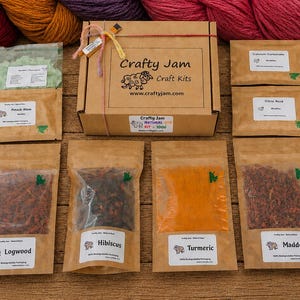 May include: A Crafty Jam craft kit box tied with string, surrounded by skeins of yarn in shades of red, orange, and purple. The kit includes several brown paper pouches containing natural dye materials labelled Logwood, Hibiscus, Turmeric, and Madder.