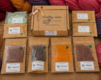 Puede incluir: Una caja de kit de manualidades Crafty Jam atada con cuerda, rodeada de madejas de hilo en tonos rojo, naranja y morado. El kit incluye varias bolsas de papel marrón que contienen materiales de tinte natural etiquetados como Logwood, Hibiscus, Turmeric y Madder.